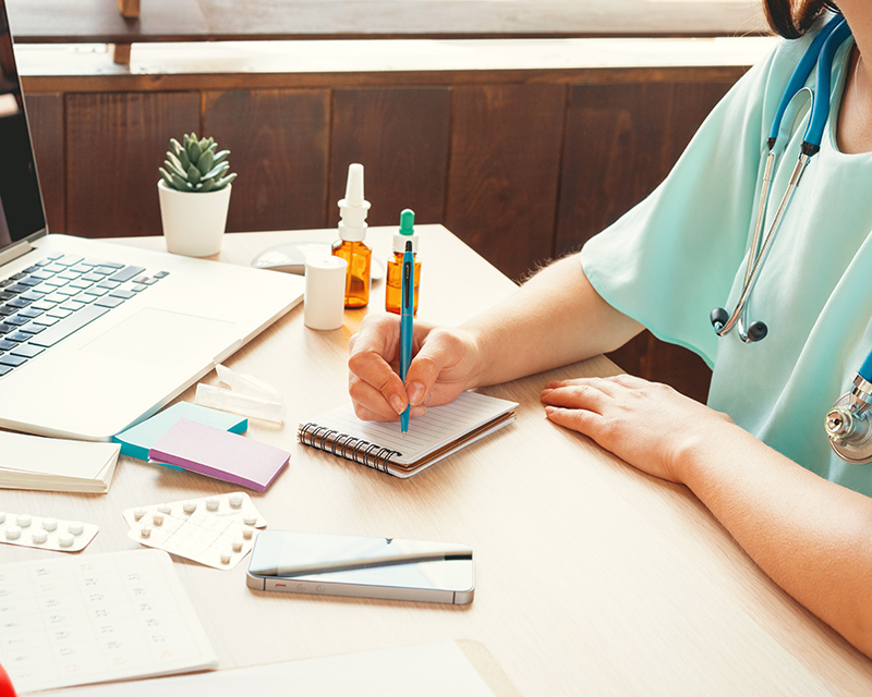A female in medical scrubs prepares paperwork at a desk with a computer and other office items