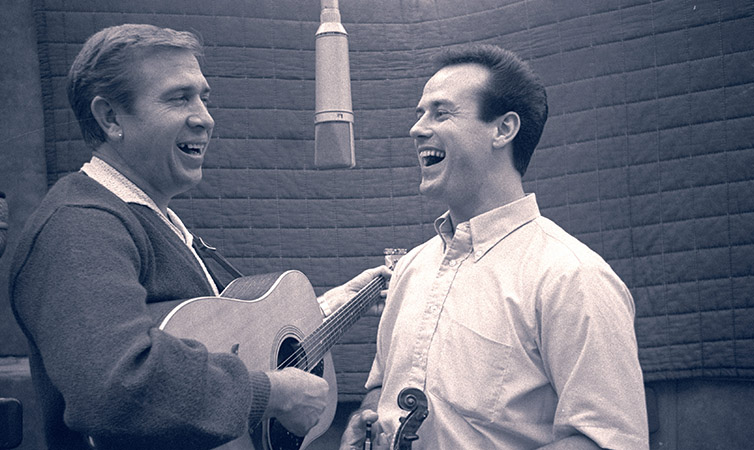 A black and white photo of Distinguished Alumnus Don Rich (left) singing into a studio microphone with Buck Owens, who is playing guitar.